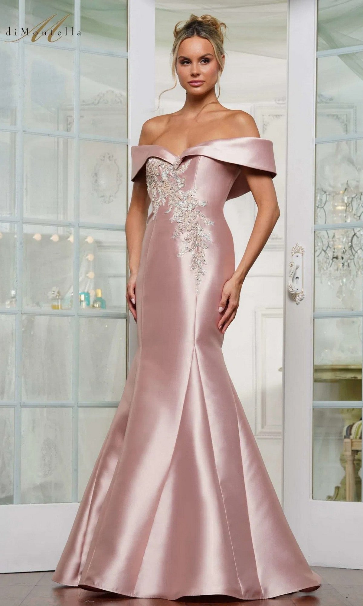 A woman stands indoors wearing the Rina Di Montella RD3257 long formal dress in blush, featuring an off-the-shoulder mermaid silhouette with beaded floral embroidery and silver embellishments on the bodice.
